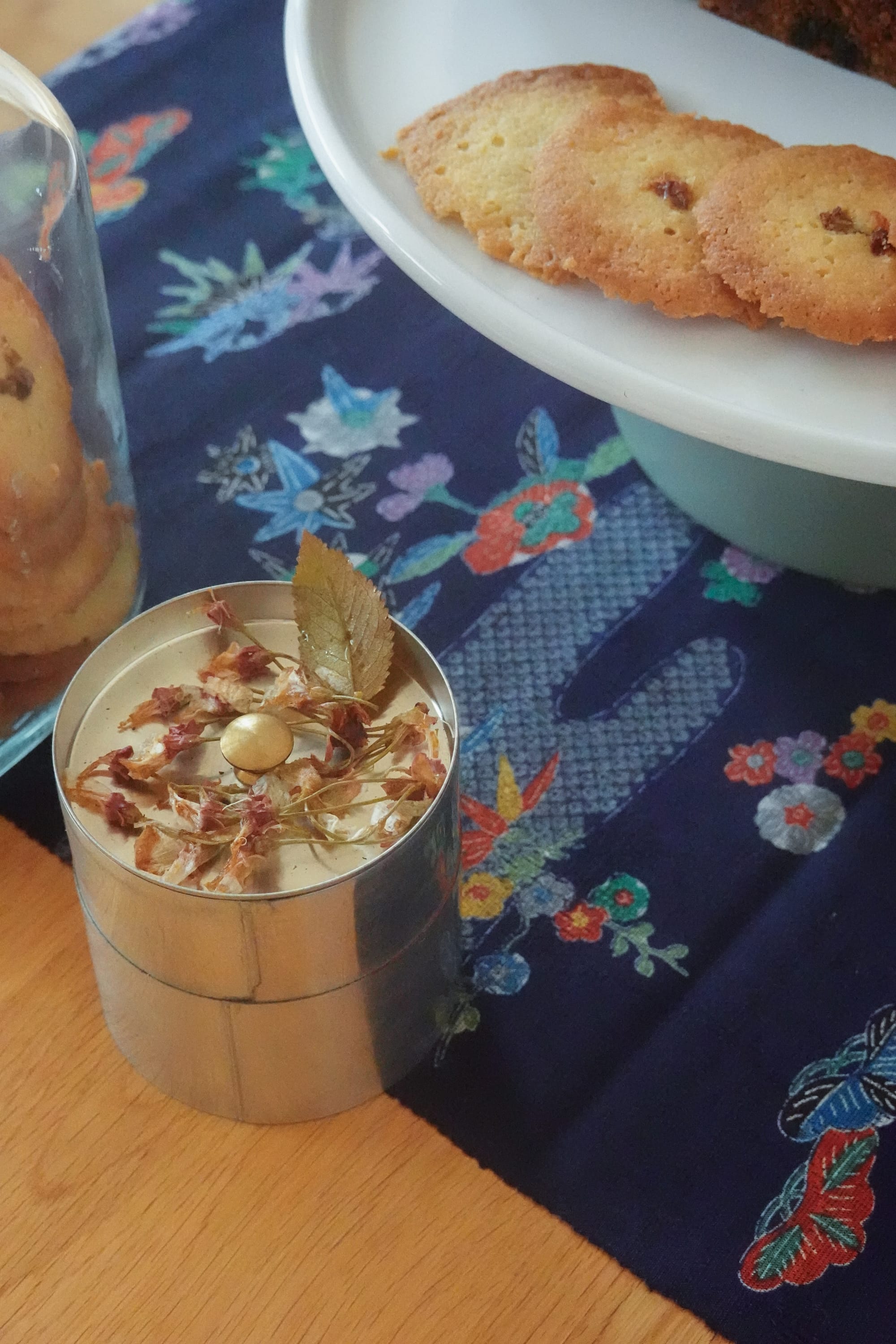 a close-up of dried cherry blossom flowers lying on a tea canister, next to tray of cookies made with the flowers