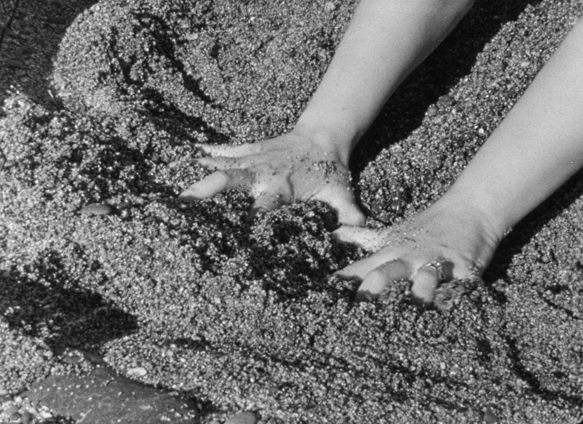 a pair of hands, with the left one wearing a ring, sink into sand with their gestures suggesting an attempt to grasp.