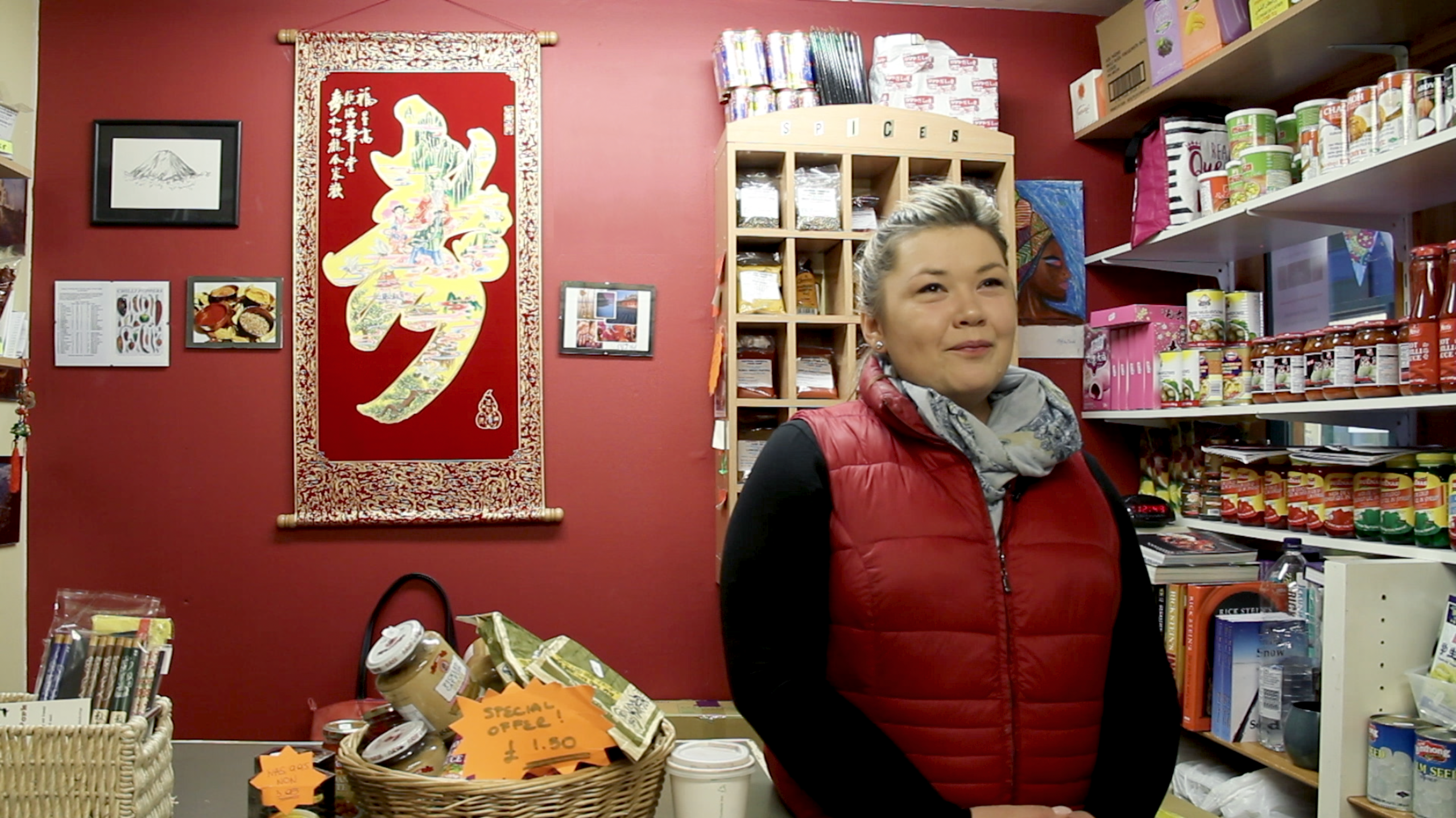 a middle aged woman with grey hair and of asian descent is wearing black long sleeve and red vest. She stands in her shop filled with asian food products with a red interior and filled shelves.