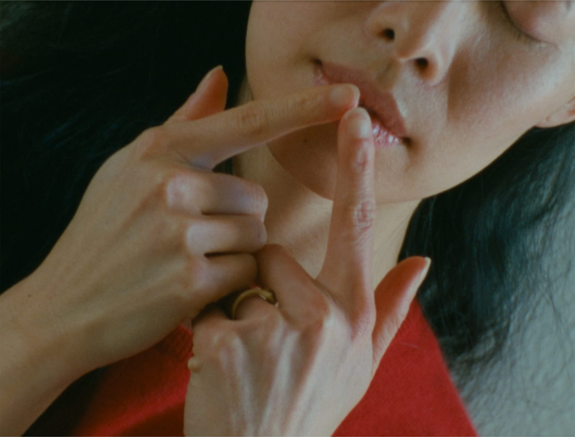 A close up of Ali, an asian woman in her thirties, wearing a red sweater with her hair down. She points to her mouth with both index finger, with her eyes closed. 
