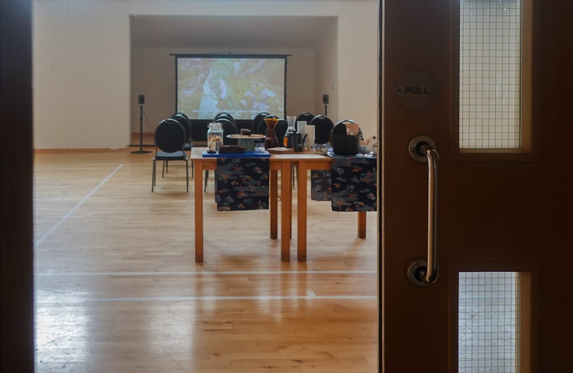 a distant view of the event's setup from a doorway, showing a projection screen at the end with a pair of speakers, lines of chairs, and two tables with clothes hosting snacks and drinks.