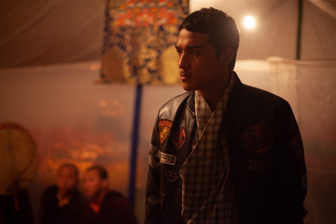 A man wearing a black jacket with patches stands indoors with two seated people blurred in the background under warm lighting.